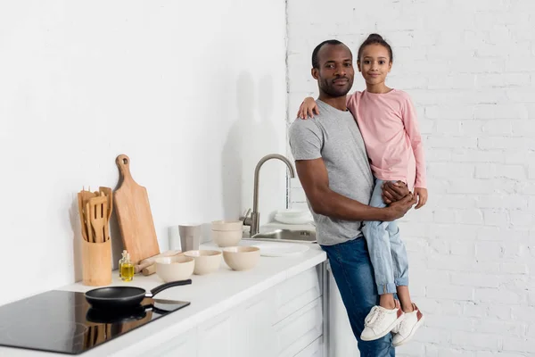 Glücklicher afrikanisch-amerikanischer Vater hält seine Tochter in der Küche und schaut in die Kamera — Stockfoto