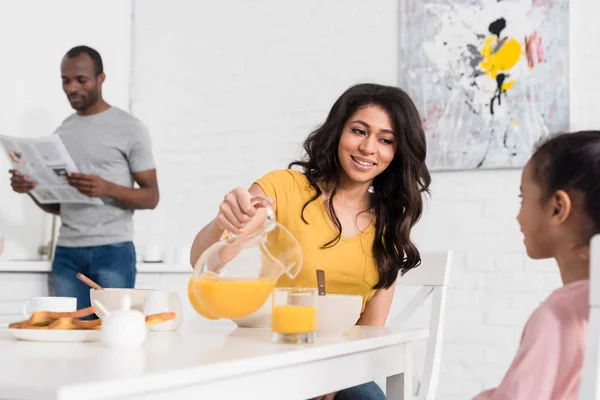 Mother pouring orange juice for daughter on breakfast while father reading newspaper blurred on background — Stock Photo