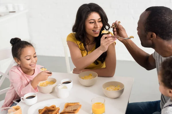 Wife and husband feeding each other while having breakfast with kids — Stock Photo