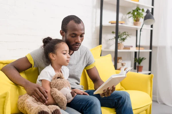 Padre guapo y pequeña hija leyendo libro juntos en casa - foto de stock