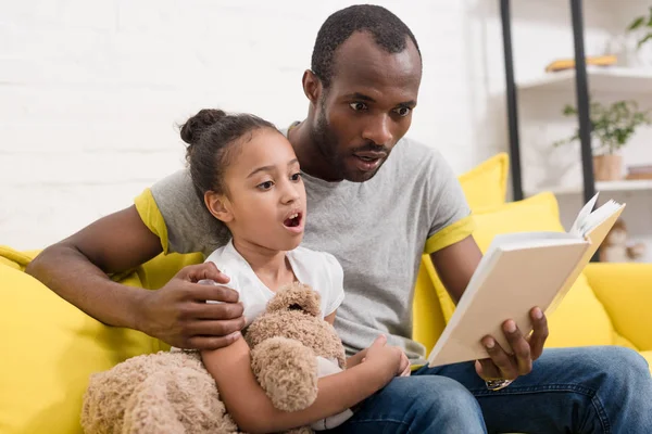 Conmocionado padre e hija leyendo el libro juntos en casa - foto de stock