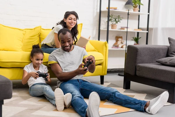 Feliz padre e hija jugando videojuegos mientras madre sentada en el sofá - foto de stock