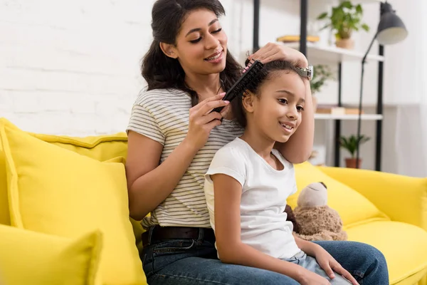 Madre peinando hermosas hijas cabello en el sofá en casa - foto de stock