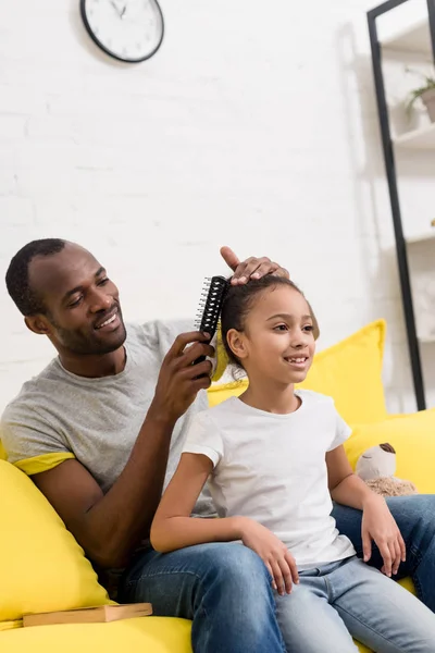 Father combing hair of daughter while sitting behind her on couch — Stock Photo