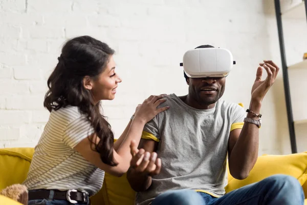 Smiling african american woman sitting near expressive boyfriend while he wearing virtual reality headset on couch at home — Stock Photo