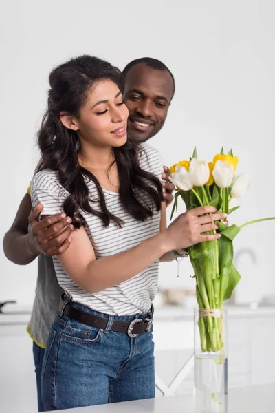 Африканская американка кладёт букет цветов в вазу, а её парень обнимает её сзади. — стоковое фото