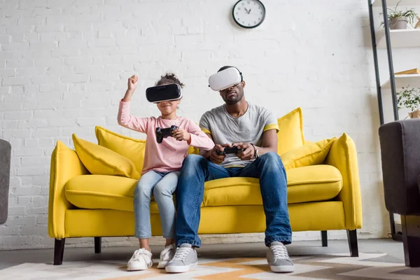 Young father and daughter in vr headsets playing video games on couch at home — Stock Photo