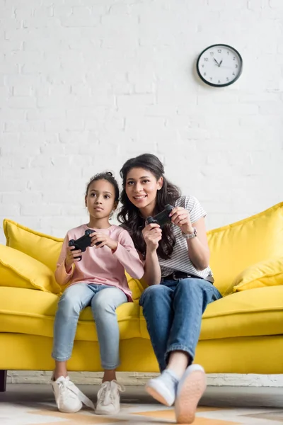 Emocional joven madre e hija jugando videojuegos en casa en el sofá - foto de stock