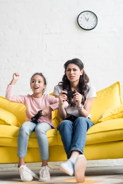 Madre e hija emocionales jugando videojuegos en casa en el sofá - foto de stock