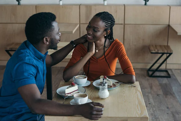 Feliz pareja afroamericana sentada en la mesa con tazas de café y postres en la cafetería - foto de stock