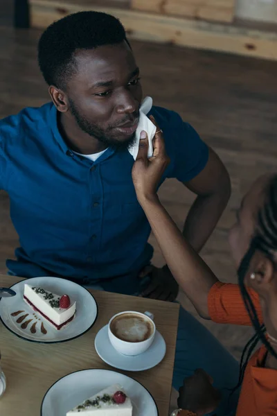 Vue grand angle de femme afro-américaine prudente nettoyage petit ami visage avec serviette à table dans le café — Photo de stock