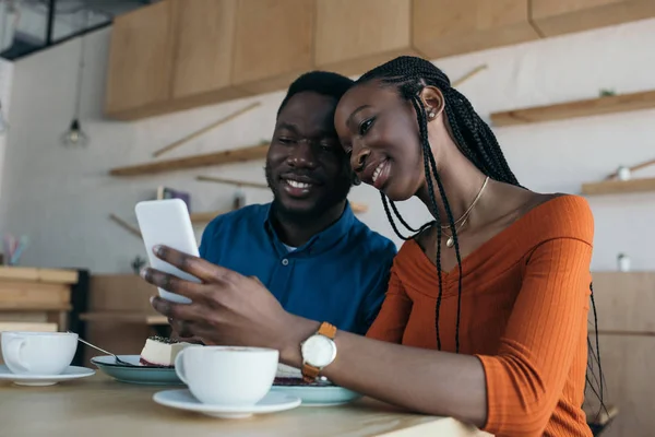 Alegre pareja afroamericana utilizando smartphone juntos en la cafetería - foto de stock