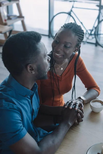 Усміхнена афро-американська пара тримає руки і дивиться один на одного на романтичне побачення в кав'ярні — стокове фото