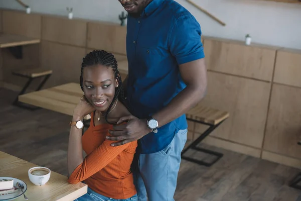 Vista parcial del hombre afroamericano abrazando a su novia sonriente en una cita romántica en una cafetería - foto de stock