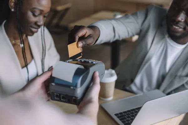 Selektiver Fokus eines afrikanisch-amerikanischen Mannes, der mit Kreditkarte bezahlt, während er Zeit mit einem Freund im Café verbringt — Stockfoto