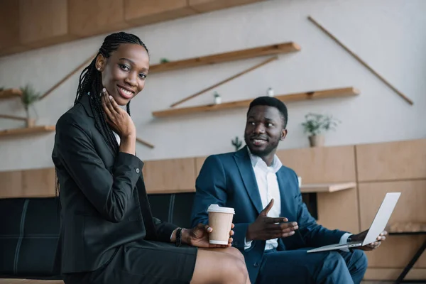 Homme d'affaires afro-américain souriant avec ordinateur portable et collègue avec café à proximité dans un café — Photo de stock