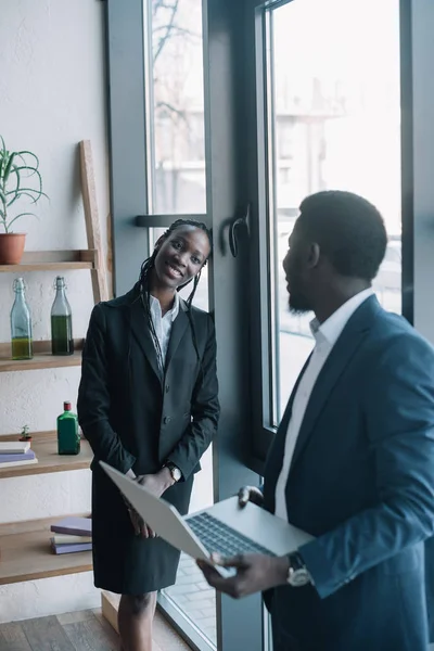 Sonriente hombre de negocios afroamericano con portátil y colega cerca en la cafetería - foto de stock