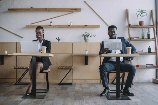 Mujer de negocios afroamericana que trabaja en el ordenador portátil mientras que el hombre de negocios que lee el periódico en la cafetería - foto de stock