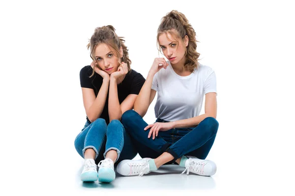 Attractive young twins sitting on floor and looking at camera on white — Stock Photo