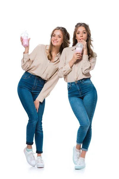 Улыбающиеся привлекательные молодые близнецы пьют молочные коктейли, изолированные на белом — стоковое фото