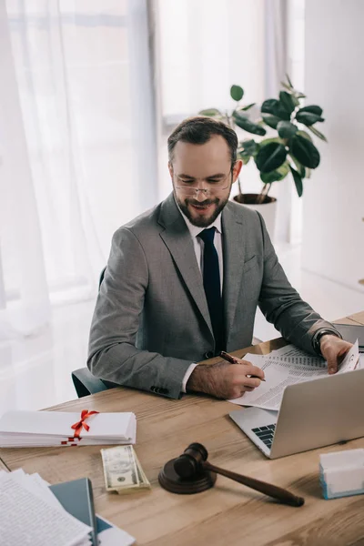 Smiling lawyer in suit looking at bribe at workplace with laptop in office — Stock Photo