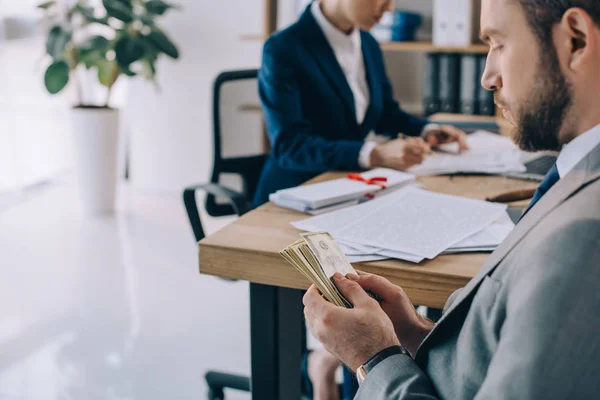 Selective focus of lawyer with dollar banknotes in hands and colleague at workplace in office — Stock Photo