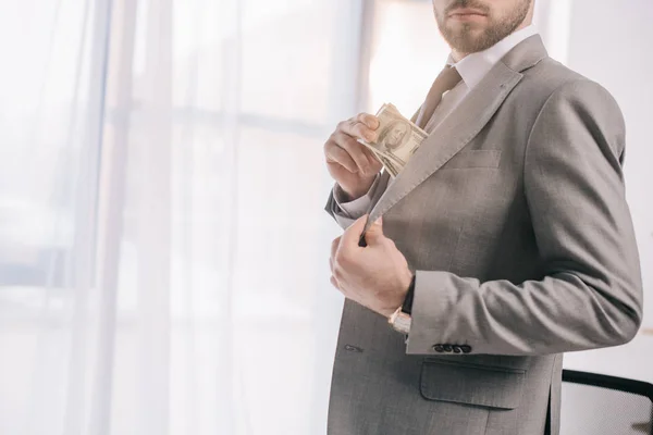Cropped shot of businessman in suit putting dollar banknotes into pocket in office — Stock Photo