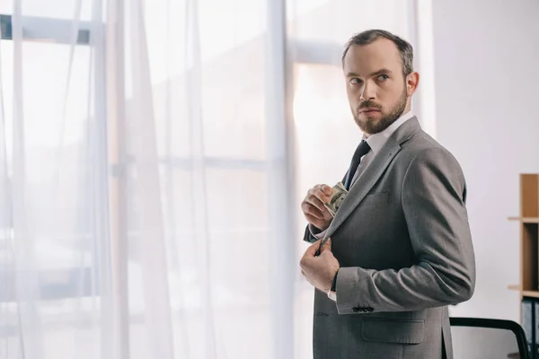 Businessman in suit putting dollar banknotes into pocket in office — Stock Photo