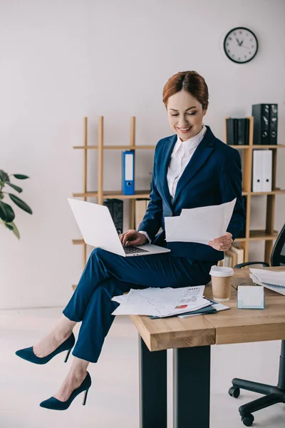 Femme d'affaires souriante avec ordinateur portable et documents assis sur la table dans le bureau — Photo de stock