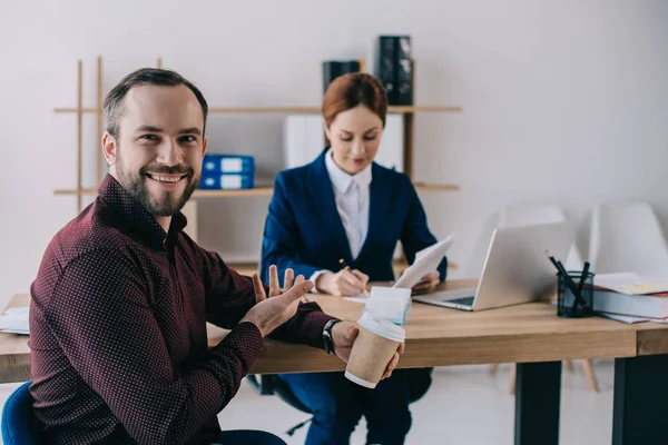 Selective focus of smiling client with coffee to go pointing at businesswoman at workplace in office — Stock Photo