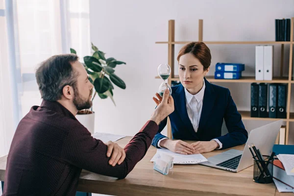 Client avec sablier et femme d'affaires sur le lieu de travail avec documents et ordinateur portable au bureau — Photo de stock