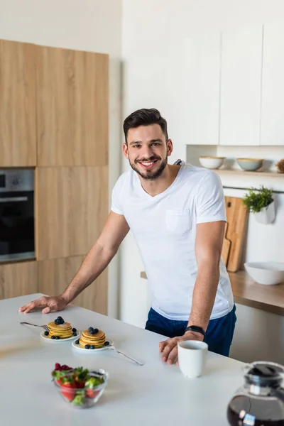 Schöner junger Mann lehnt am Küchentisch und lächelt morgens in die Kamera — Stockfoto