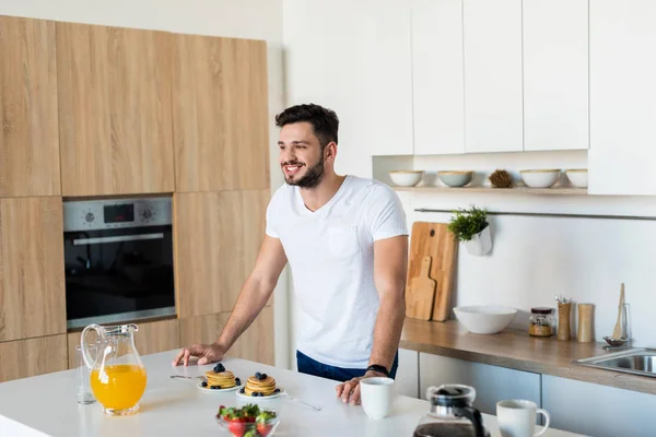 Улыбающийся молодой человек склоняется к кухонному столу с завтраком и смотрит в сторону — стоковое фото