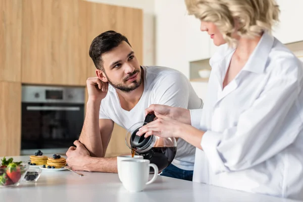 Jovem mulher derramando café e olhando para namorado bonito de manhã na cozinha — Fotografia de Stock