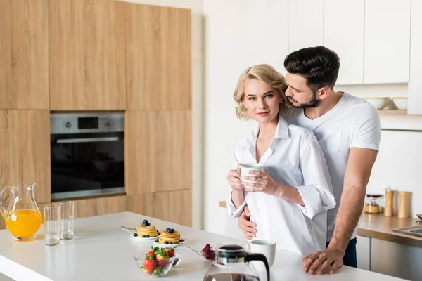 Joven abrazando novia sosteniendo taza de café y mirando a la cámara en la cocina - foto de stock