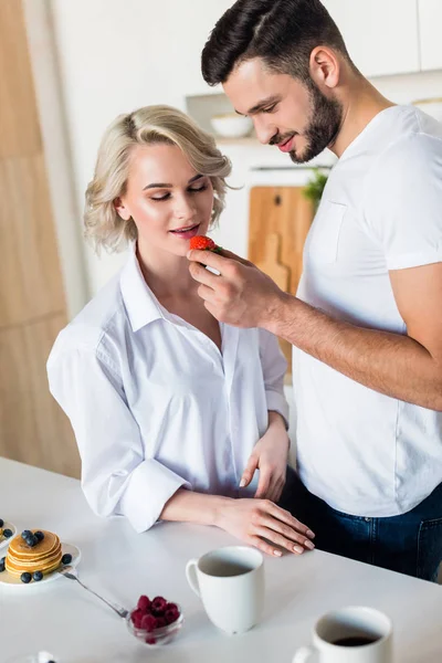 Smiling young man feeding sexy girlfriend with strawberry at morning — Stock Photo