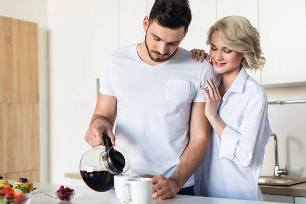 Lächelnde junge Frau umarmt schönen Freund, während er Kaffee in der Küche einschenkt — Stockfoto