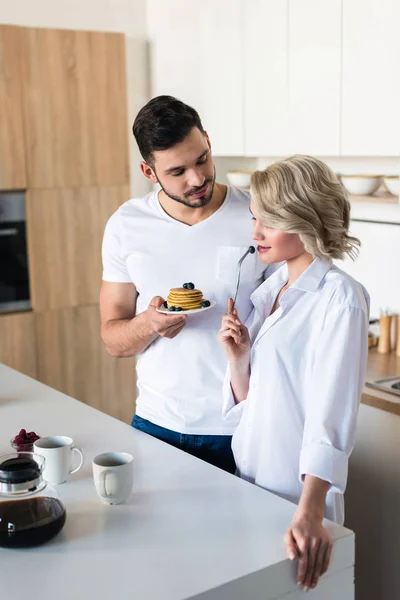Jovem homem segurando panquecas e olhando para namorada sexy na cozinha — Fotografia de Stock
