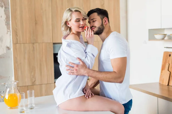 Vue latérale d'un jeune homme souriant embrassant sa copine sexy mangeant des fraises dans la cuisine — Photo de stock