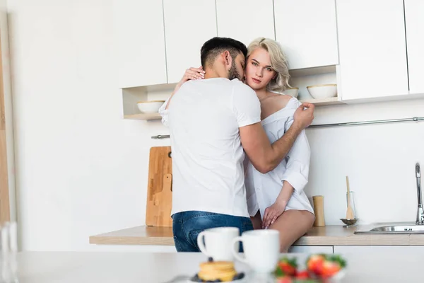 Jeune homme étreignant et embrassant petite amie sexy, fille regardant la caméra dans la cuisine — Photo de stock