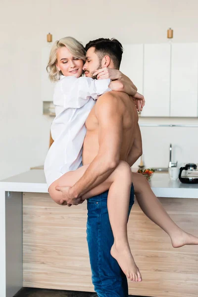 Beau torse nu jeune homme portant et étreignant petite amie sexy dans la cuisine — Photo de stock