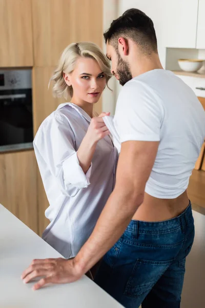 Seductora mujer joven mirando a la cámara y quitándose la camiseta de novio guapo en la cocina - foto de stock