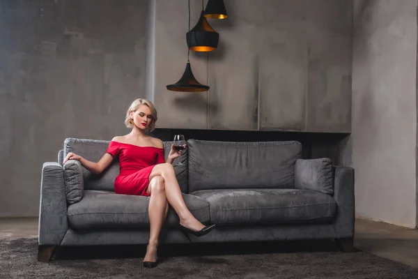 Belle femme blonde en robe rouge assise sur canapé et buvant du vin — Photo de stock