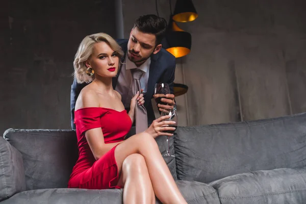 Seductive young couple drinking wine, woman holding necktie of boyfriend and looking at camera — Stock Photo