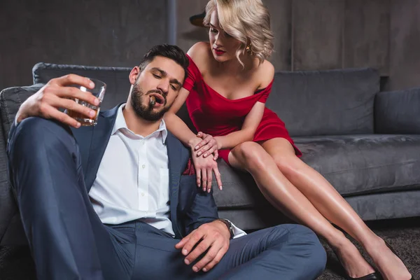 Seductive young woman in red dress looking at handsome man in suit smoking cigar and drinking whiskey — Stock Photo