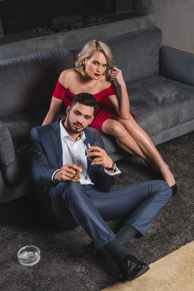 Bel homme en costume tenant cigare et verre de whisky tout en passant du temps avec une femme séduisante en robe rouge — Photo de stock