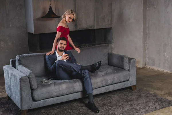 Seductive young woman in red dress looking at handsome boyfriend in suit smoking cigar and looking at camera — Stock Photo