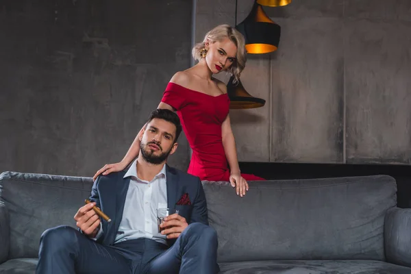 Hombre de traje sosteniendo cigarro y vaso de whisky mientras pasa tiempo con chica sexy en vestido rojo - foto de stock