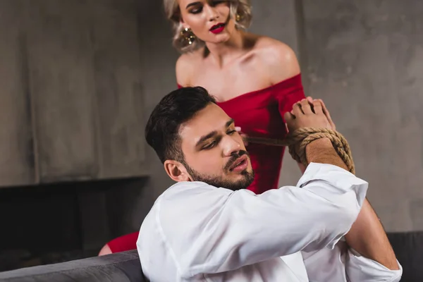 Sensual jovem mulher em vestido vermelho amarrando as mãos de bonito namorado com corda — Fotografia de Stock