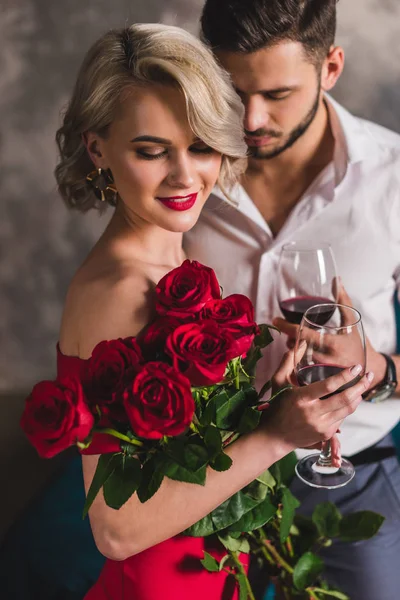 Sonriente joven en vestido rojo sosteniendo hermosas rosas y bebiendo vino con guapo novio — Stock Photo